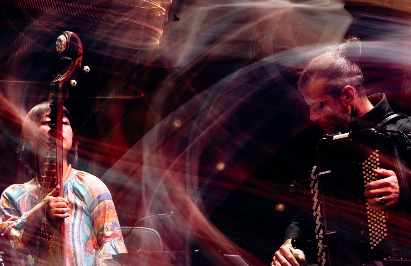 Two musicians perform on stage, one with a pipa, one with an accordion. They are surrounded by swirling light trails.