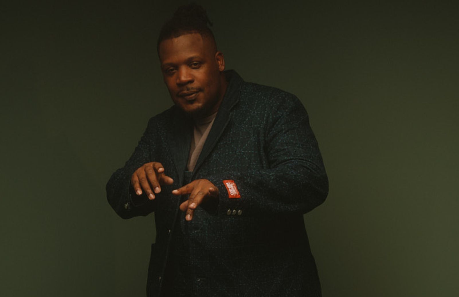 A man in a dark blazer stands against a deep green background.