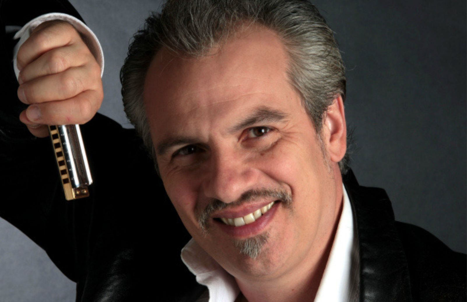 A smiling man holding a harmonica against a gray background.