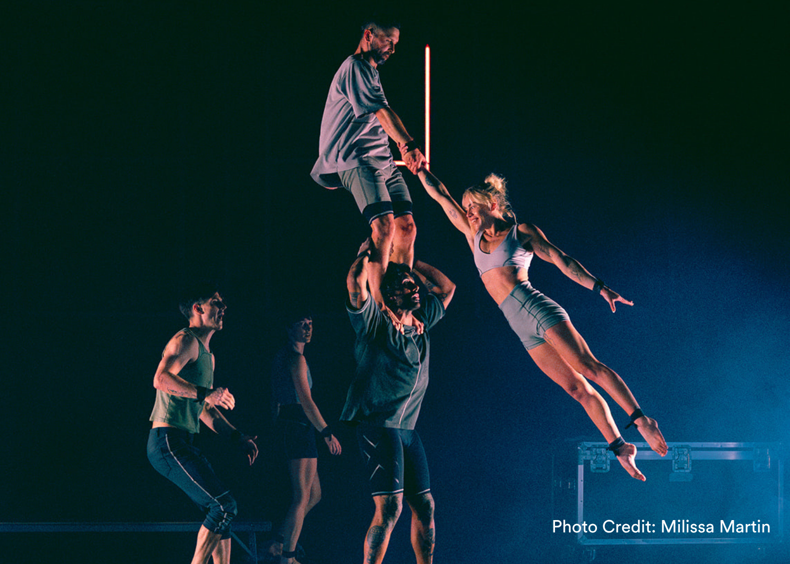An acrobat is extended horizontally in midair, held by a performer atop a human tower, as others watch from a darkened stage. (Photo Credit: Milissa Martin)