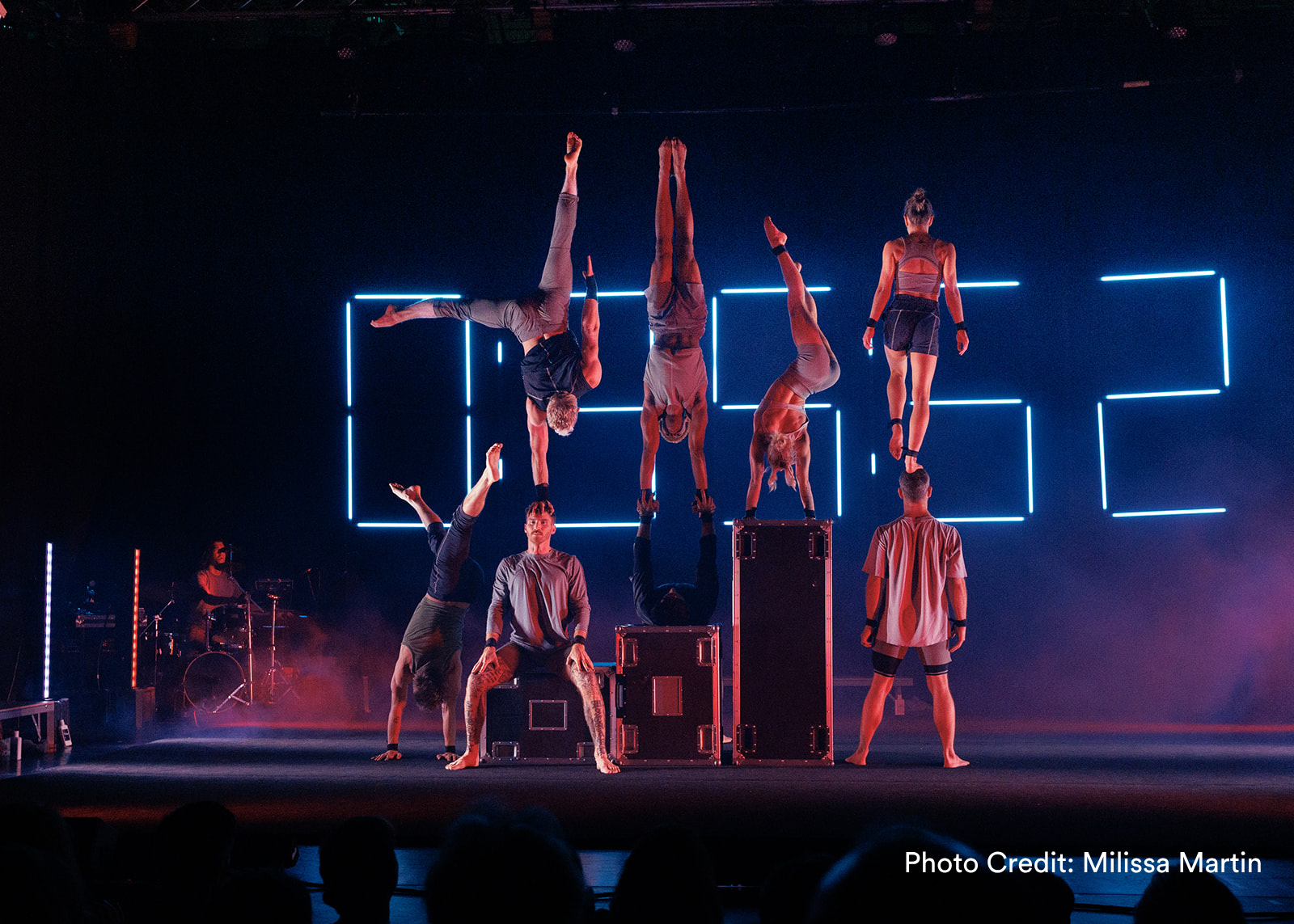 Acrobatic performers hold handstands on cases and each other against glowing neon clock digits on a dramatic stage. (Photo Credit: Milissa Martin)