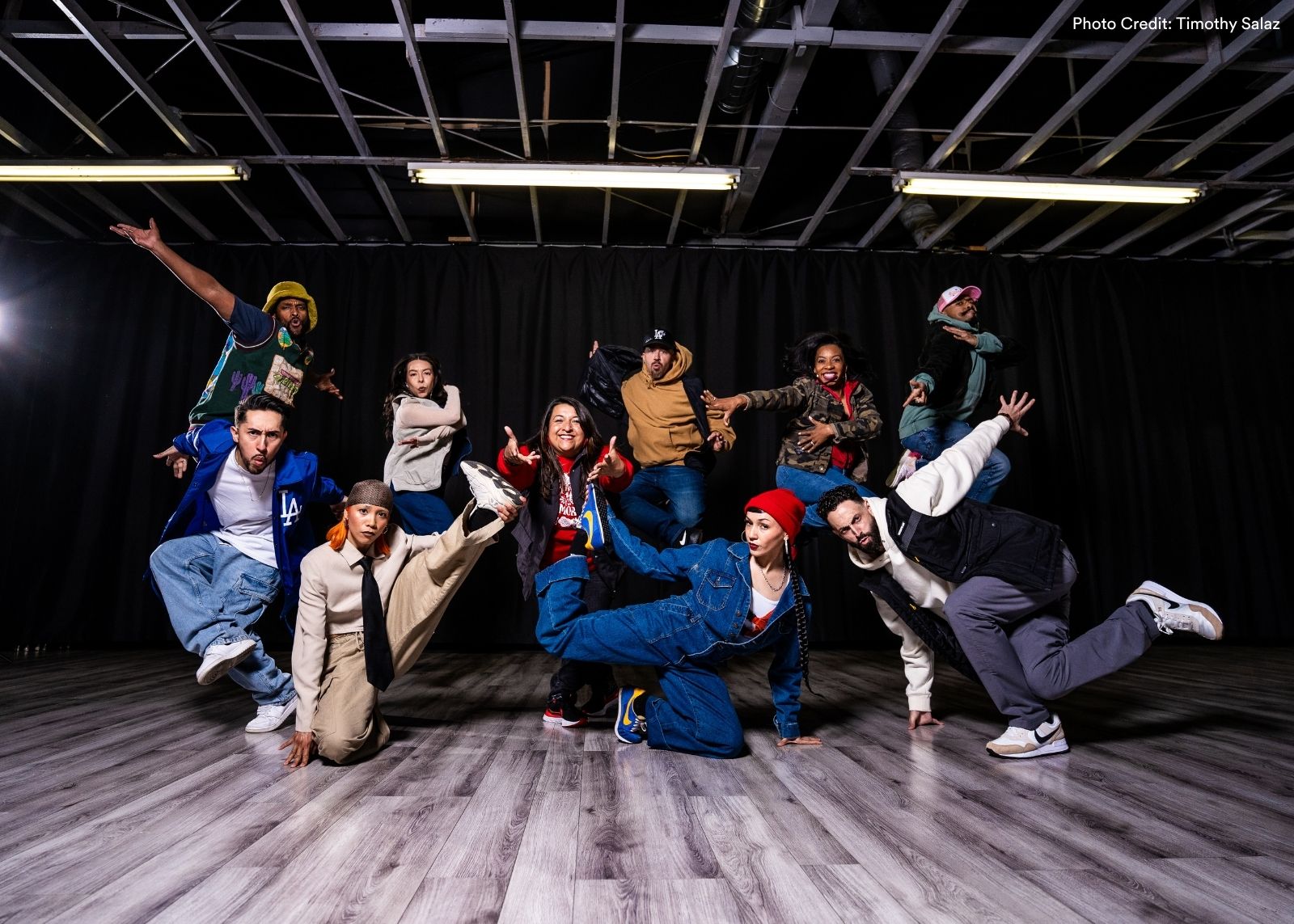 The full Versa-Style Dance Company ensemble strikes dynamic poses in a studio, some jumping and others low to the ground, lit dramatically against a black curtain backdrop. Photo: Timothy Salaz.