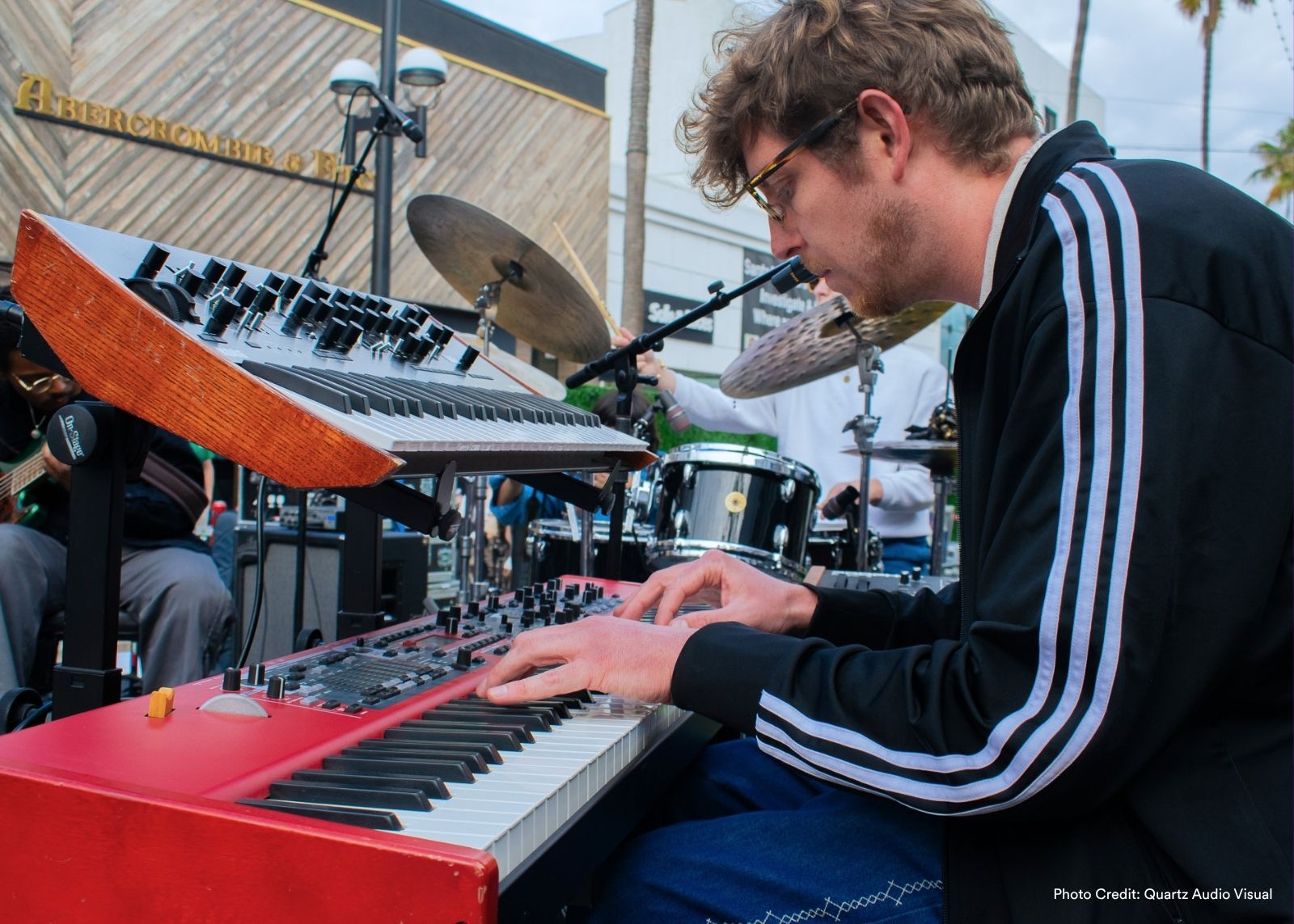 A keyboardist in a black track jacket plays a red keyboard on an outdoor stage.