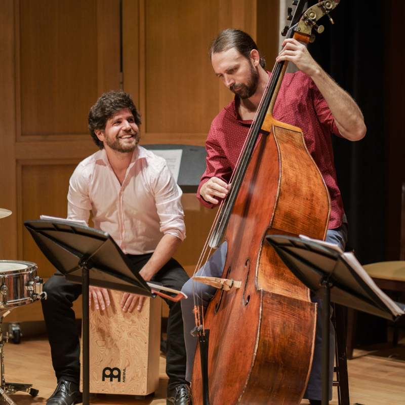 Two musicians smile while playing cajon and double bass in a wood-paneled concert hall.