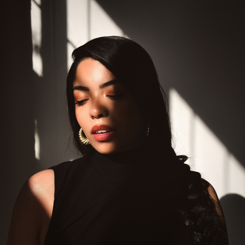 A woman with eyes closed tilts her head downward in dramatic shadow-play lighting, wearing a black top and gold hoop earrings. Photo: Shervin Lainez.