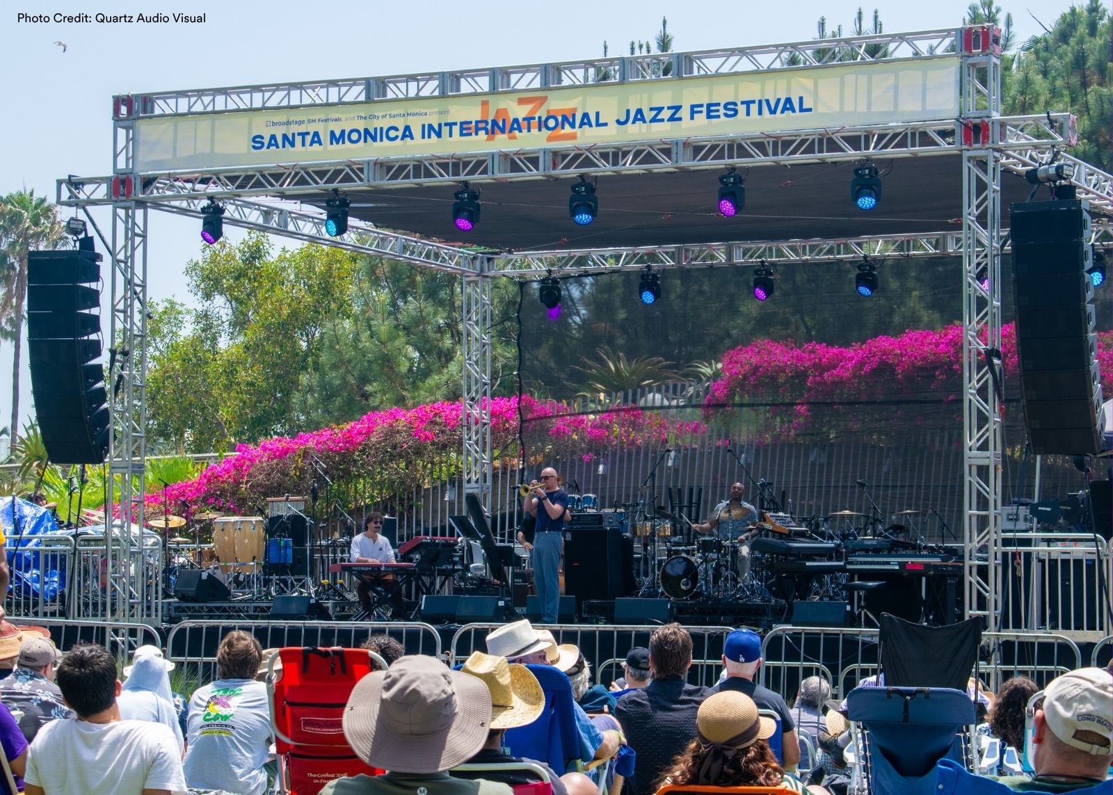 The Santa Monica International Jazz Festival main stage at Tongva Park, framed by truss and festival signage, with a trumpet player center stage and a crowd seated in the sunshine.