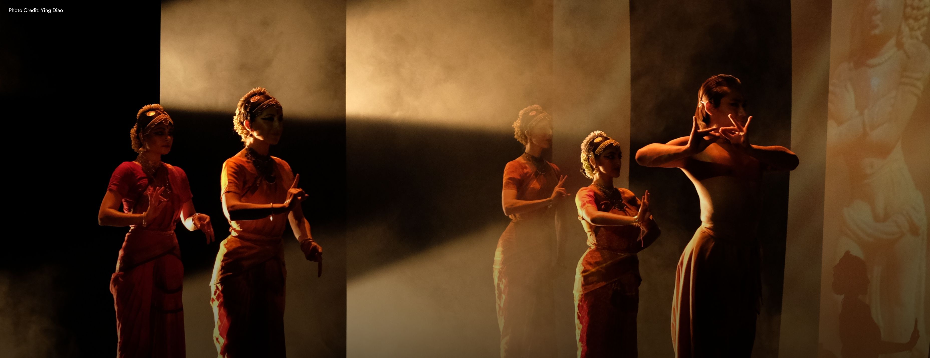 Three dancers in traditional South Asian attire perform on a dramatically lit stage with warm amber light and projected imagery in the background. Photo: Ying Gao.