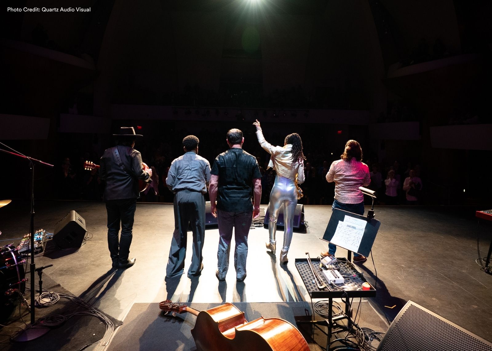 Five musicians take a bow on a concert stage, facing an applauding audience, with instruments including a cello and guitar visible in the foreground.