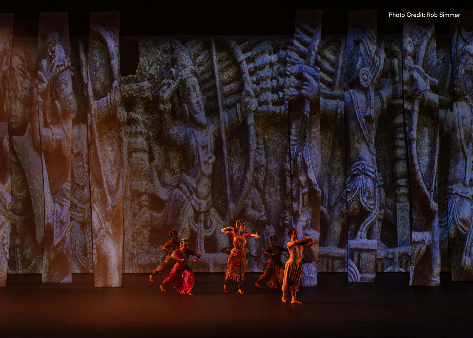 Four classical Indian dancers move across a vast stage, next to towering projected images of temple carvings. Photo: Rob Simmer.