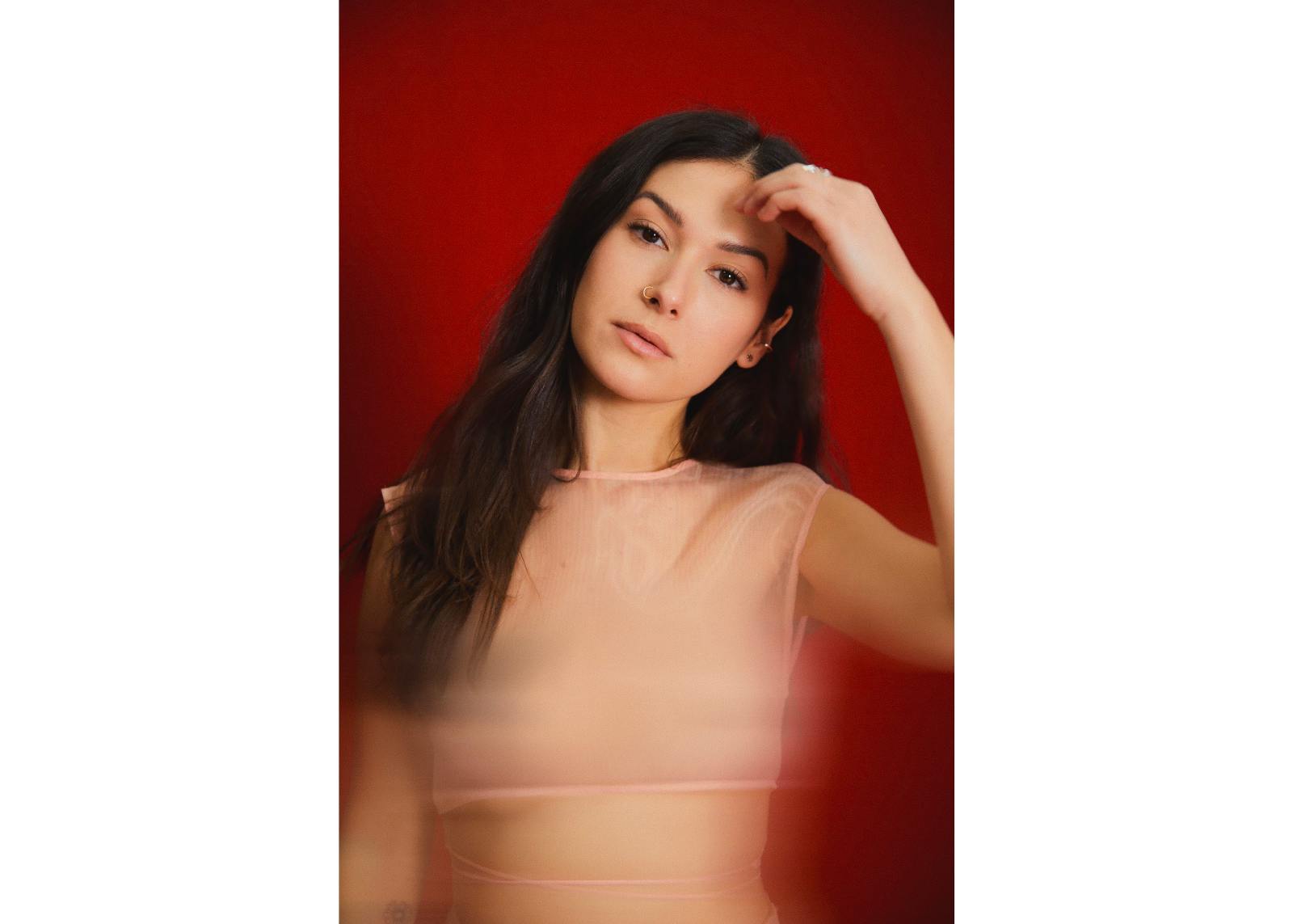 A woman with long dark hair gazes at the camera with one hand raised to her forehead, wearing a sheer pink top against a deep red background.