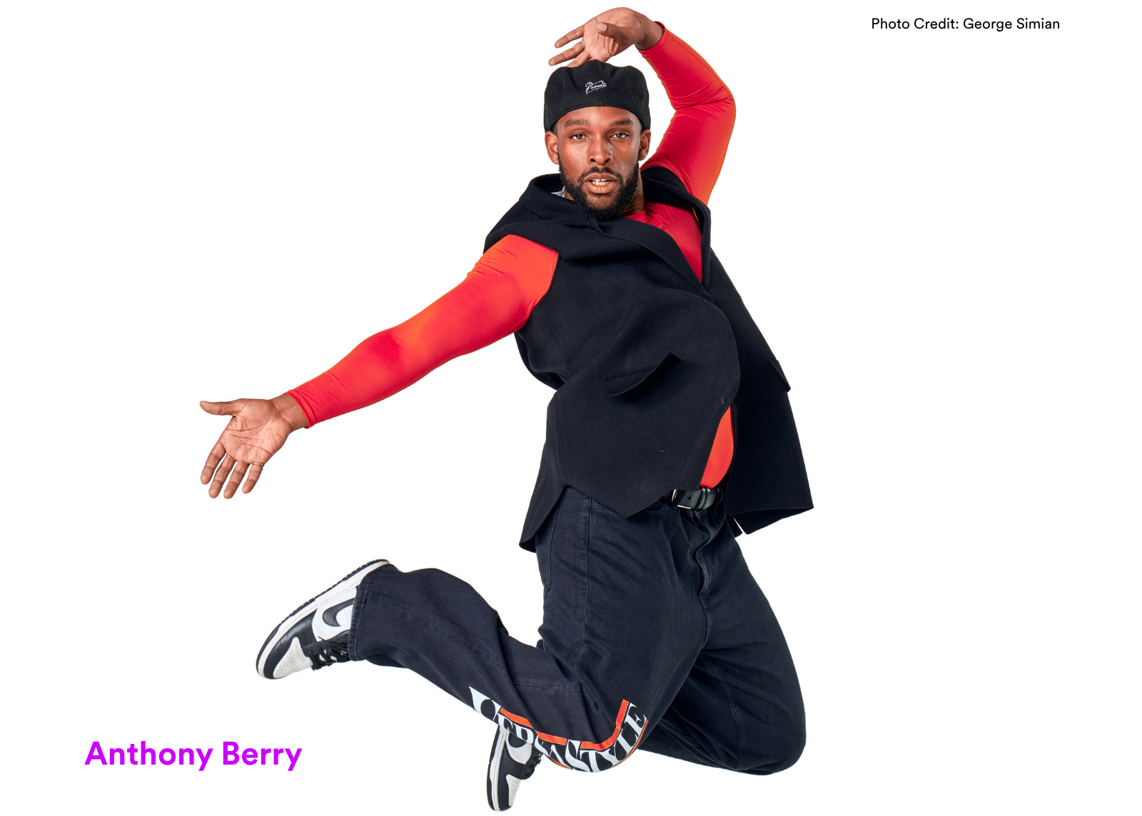 Dancer Anthony Berry leaps mid-air in a red long-sleeve shirt and black cargo pants, one arm raised and the other extended, against a white background. Photo: George Simian.