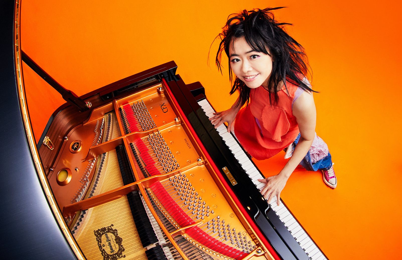 A pianist in a red dress smiles from inside an open grand piano, shot from above against a bright orange background.