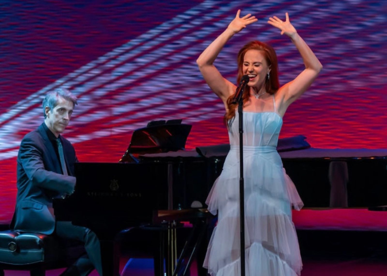 Singer in a light blue tiered dress performs at a standing microphone with arms raised, accompanied by a pianist against a red and blue projection backdrop.