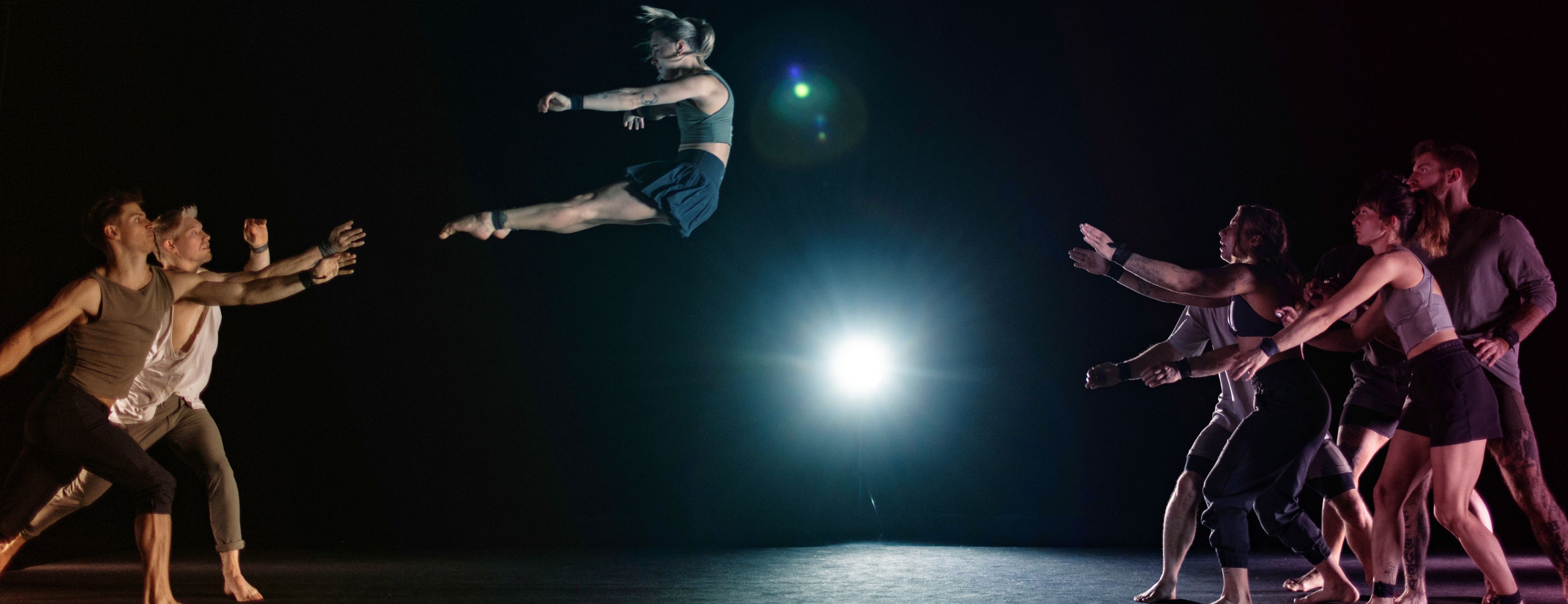 A dancer leaps high above a darkened stage as two groups of dancers reach toward her from either side.