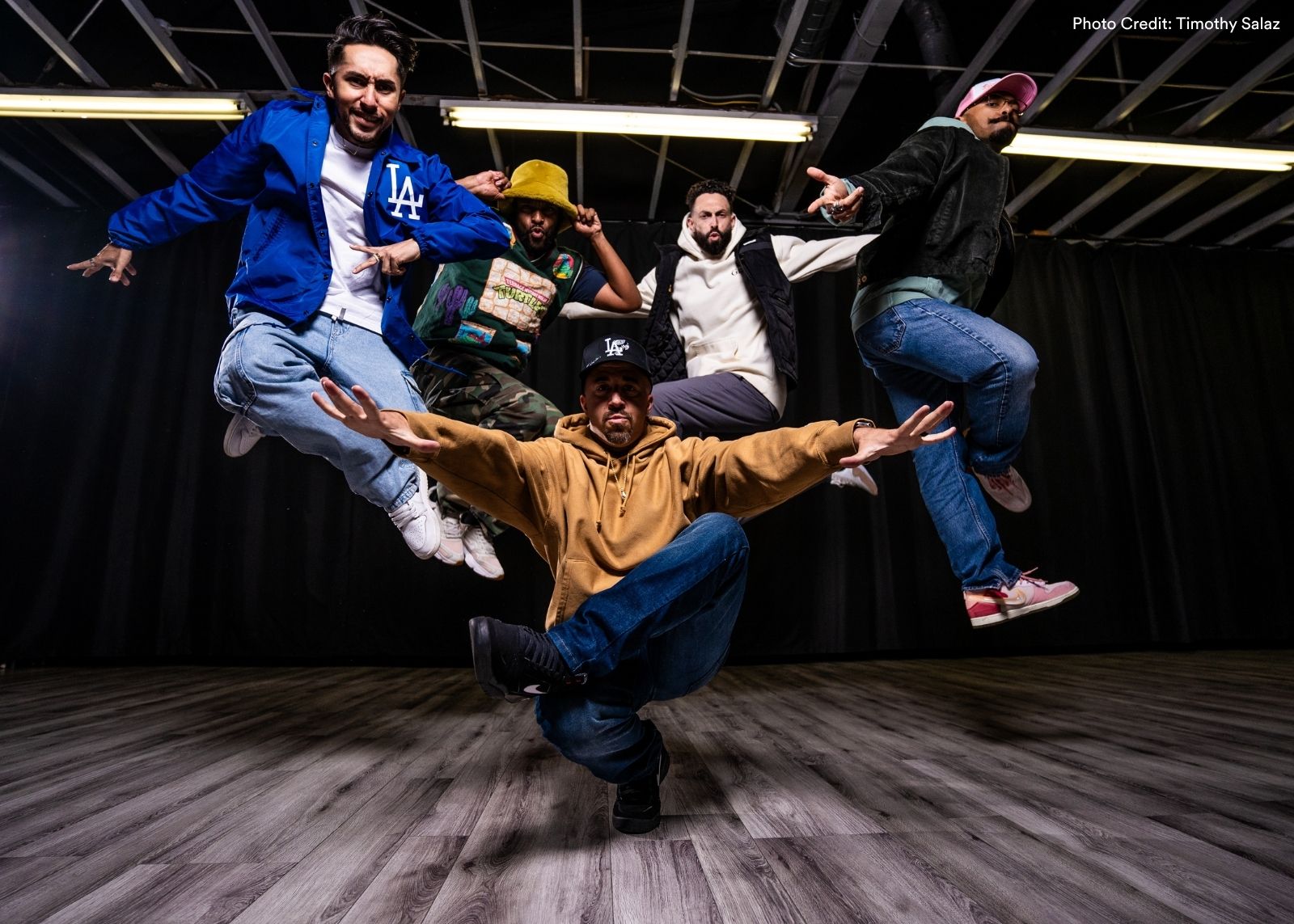 Five male dancers from Versa-Style Dance Company jump and crouch in formation in a studio, one centered low to the ground with arms wide as others leap on either side. Photo: Timothy Salaz.