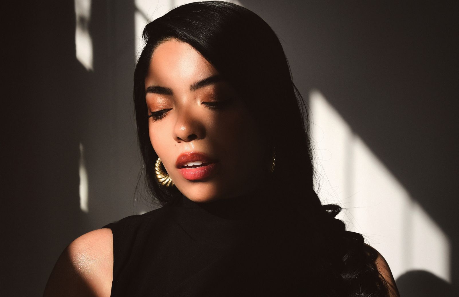 A woman in a black top with gold hoop earrings gazes downward in dramatic natural light and shadow.