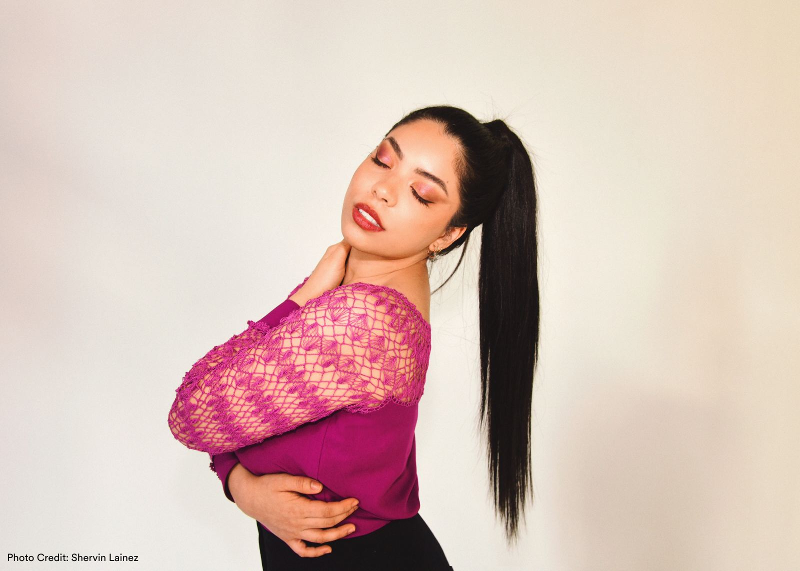 A woman with eyes closed tilts her head back, wearing a magenta lace off-shoulder top with a long sleek ponytail. Photo: Shervin Lainez.