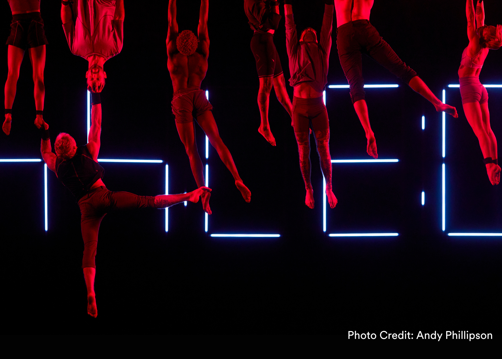 A performer is lifted to stand on one arm above four acrobats, all framed by a glowing red neon countdown clock. (Photo Credit: Andy Phillipson)