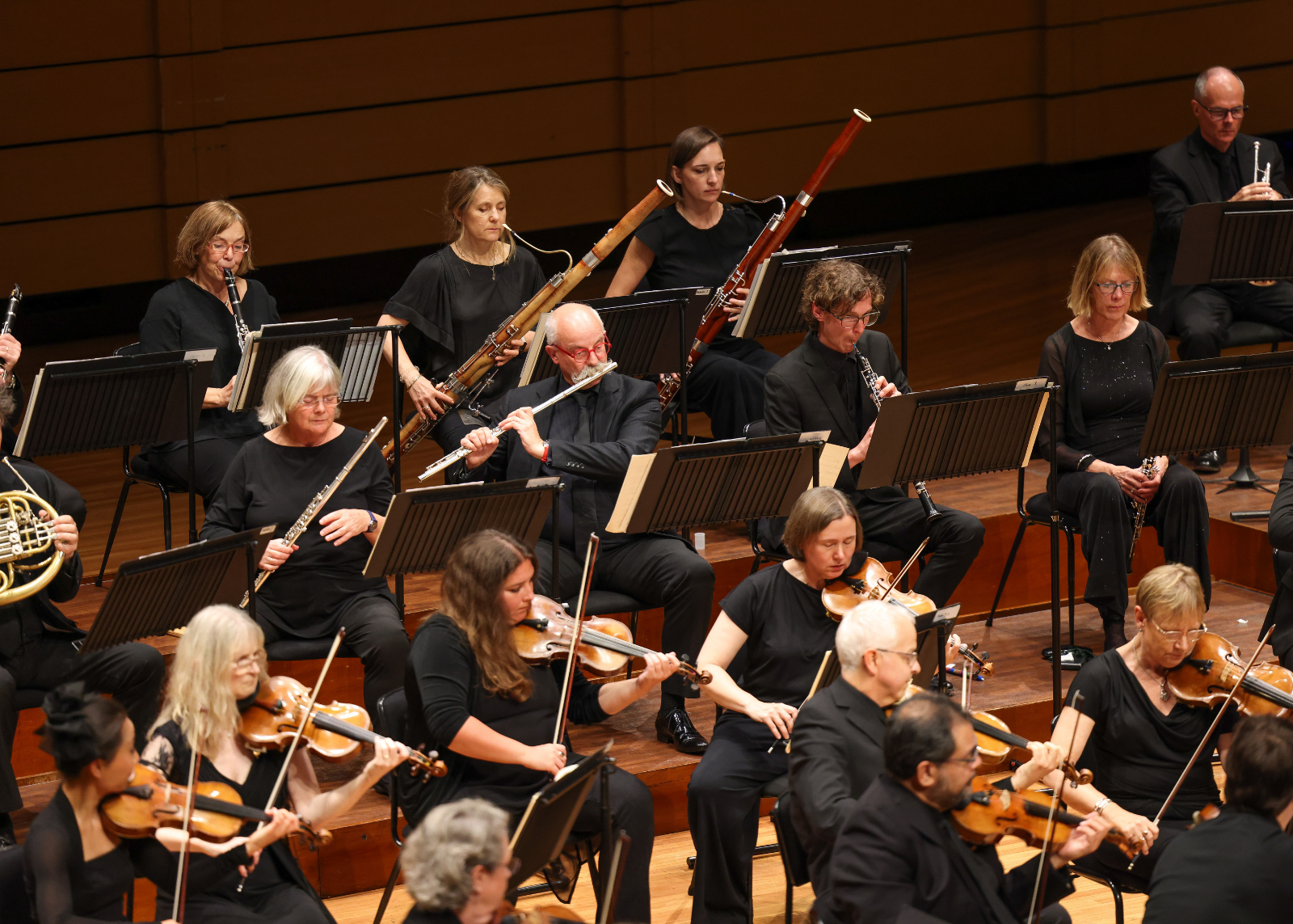 Orchestra woodwind and string sections perform on stage, with flute, bassoon, clarinet, and violin players visible in formal black attire against warm wood paneling.