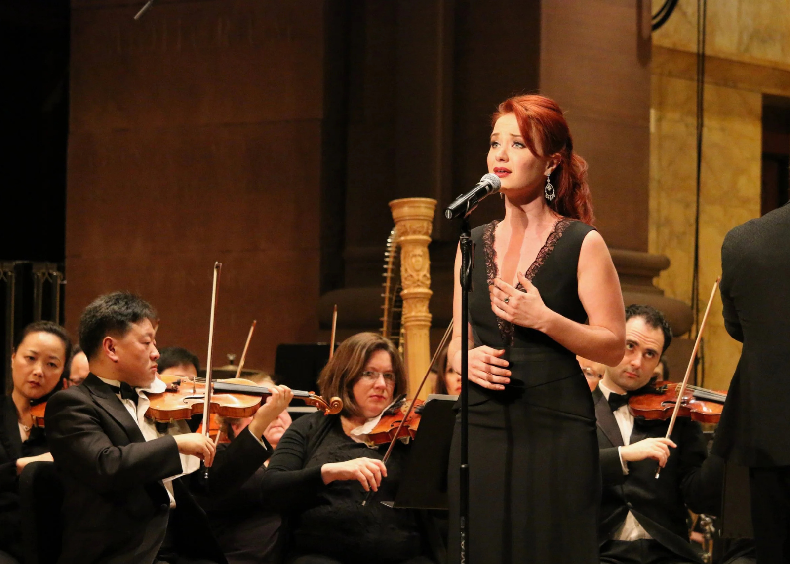 Singer in a black lace-trimmed dress performs at a standing microphone with an orchestra behind her.