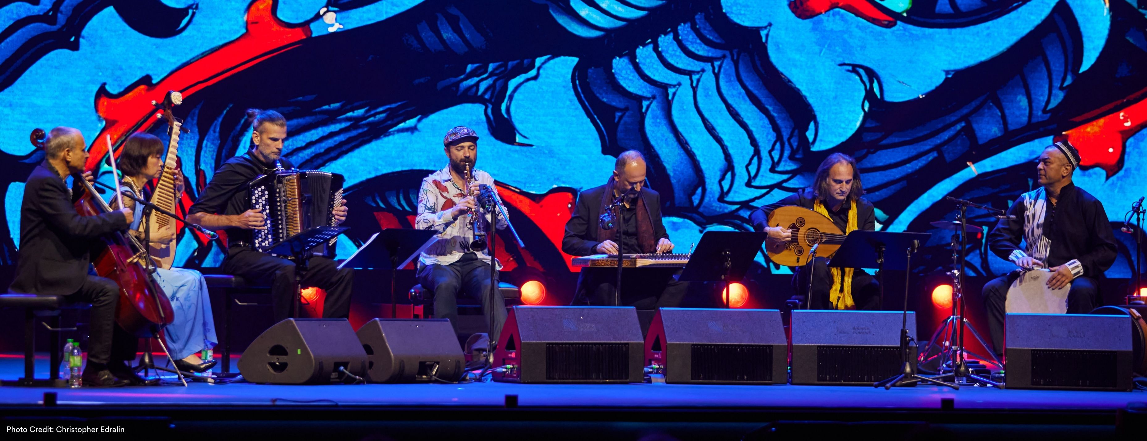Seven musicians perform on stage against a vivid blue and red painted backdrop, playing cello, pipa, accordion, saxophone, oud, and frame drum. (Photo Credit: Christopher Edralin)