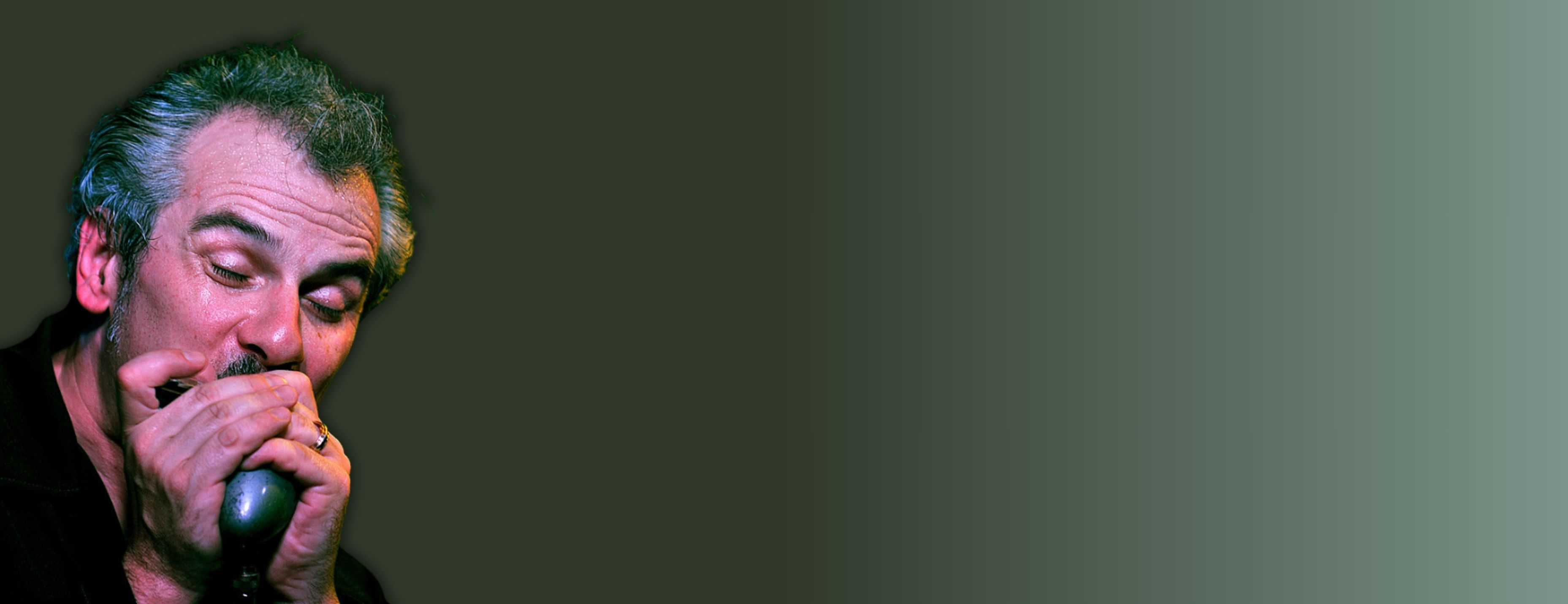 A man with white hair plays the harmonica in front of a green gradient background.