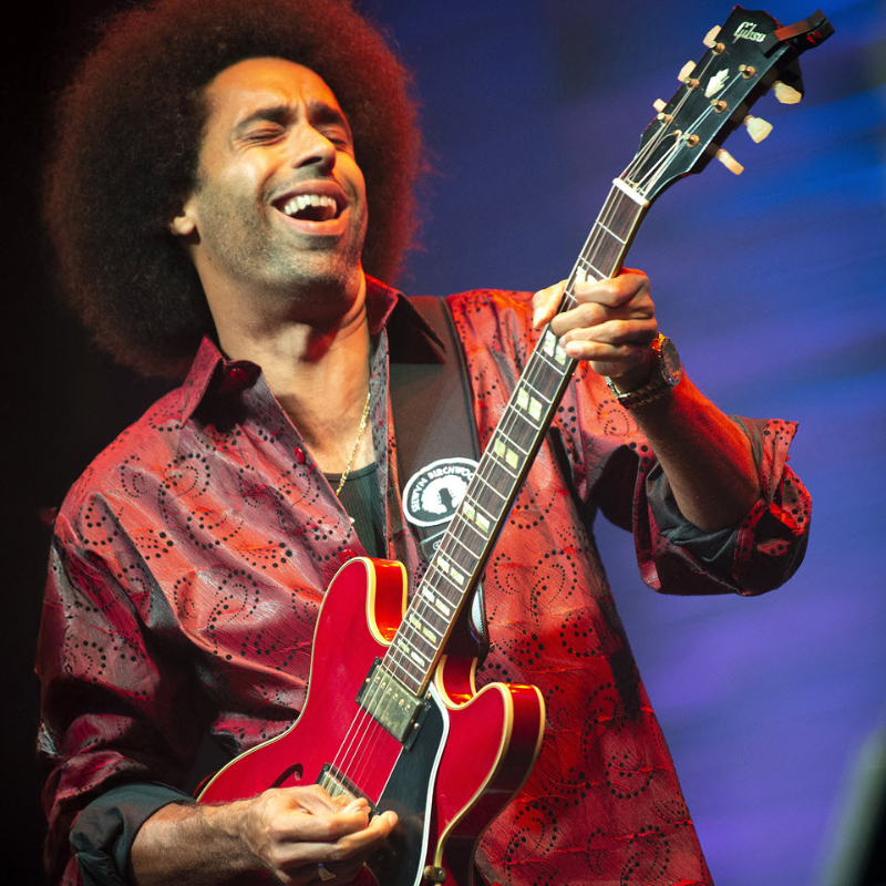 Selwyn Birchwood plays electric guitar and laughs with eyes closed under blue stage lighting, wearing a patterned shirt.