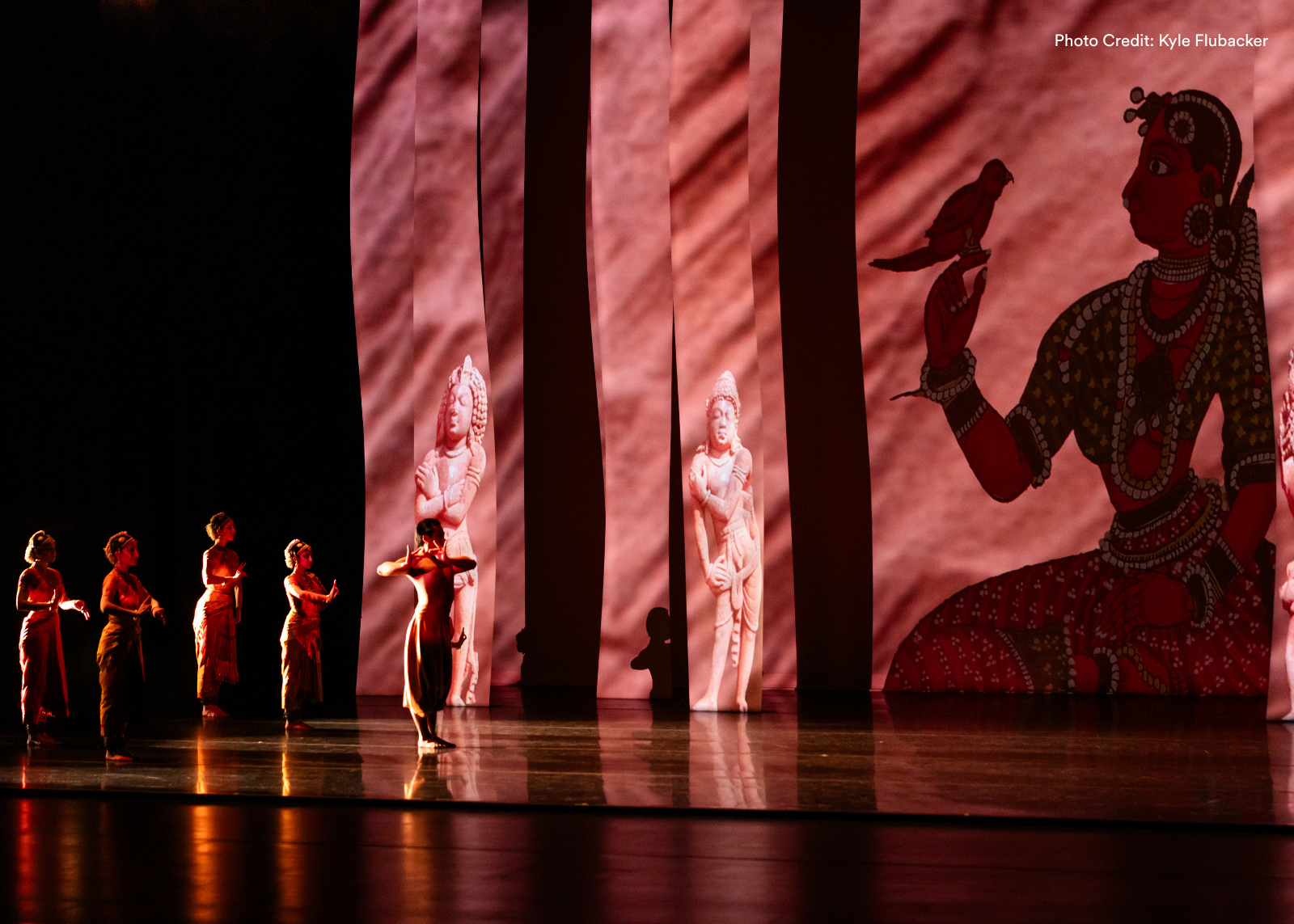 A line of classical Indian dancers performs on a darkened stage bathed in deep red light, with towering projected panels displaying sculptures and artwork. Photo: Kyle Flubacker.