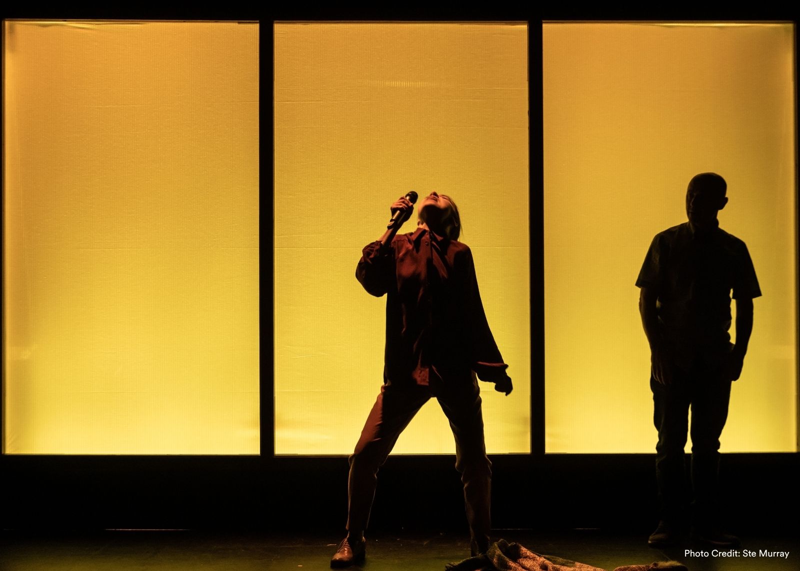 A performer sings into a microphone with head thrown back, silhouetted against three glowing golden panels, with a second person in shadow nearby.