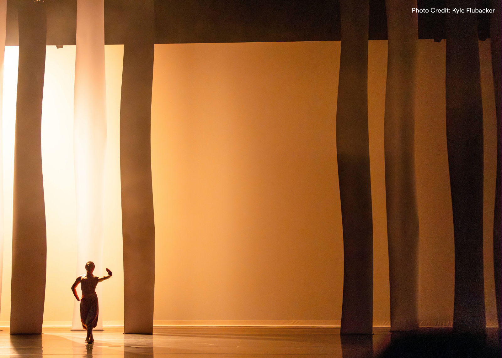 A lone dancer appears as a small silhouette between towering illuminated panels on a vast warm-lit stage. Photo: Kyle Flubacker.