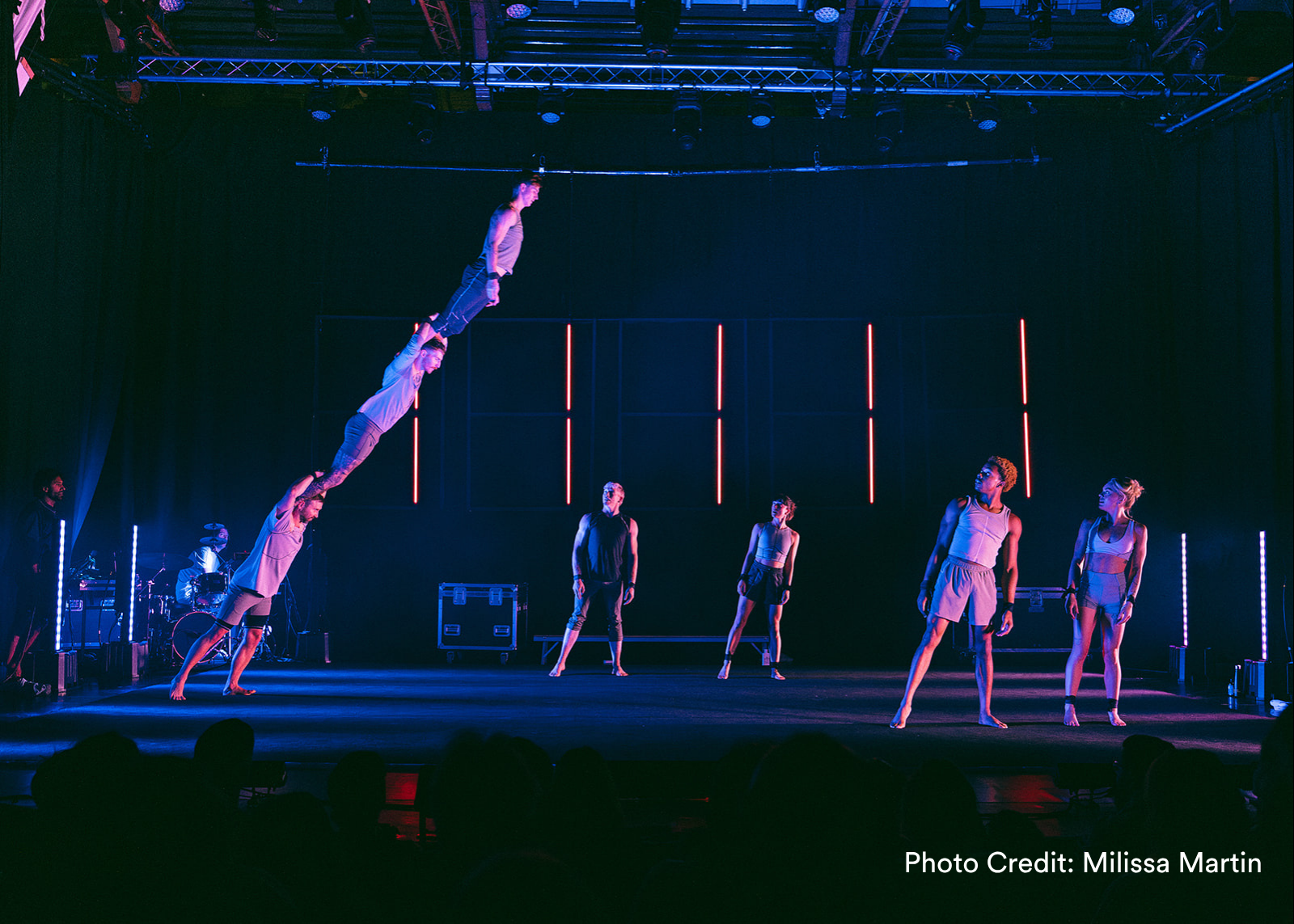 Multiple acrobats hang upside down in red light against a neon-lit backdrop, legs splayed dramatically. (Photo Credit: Andy Phillipson)