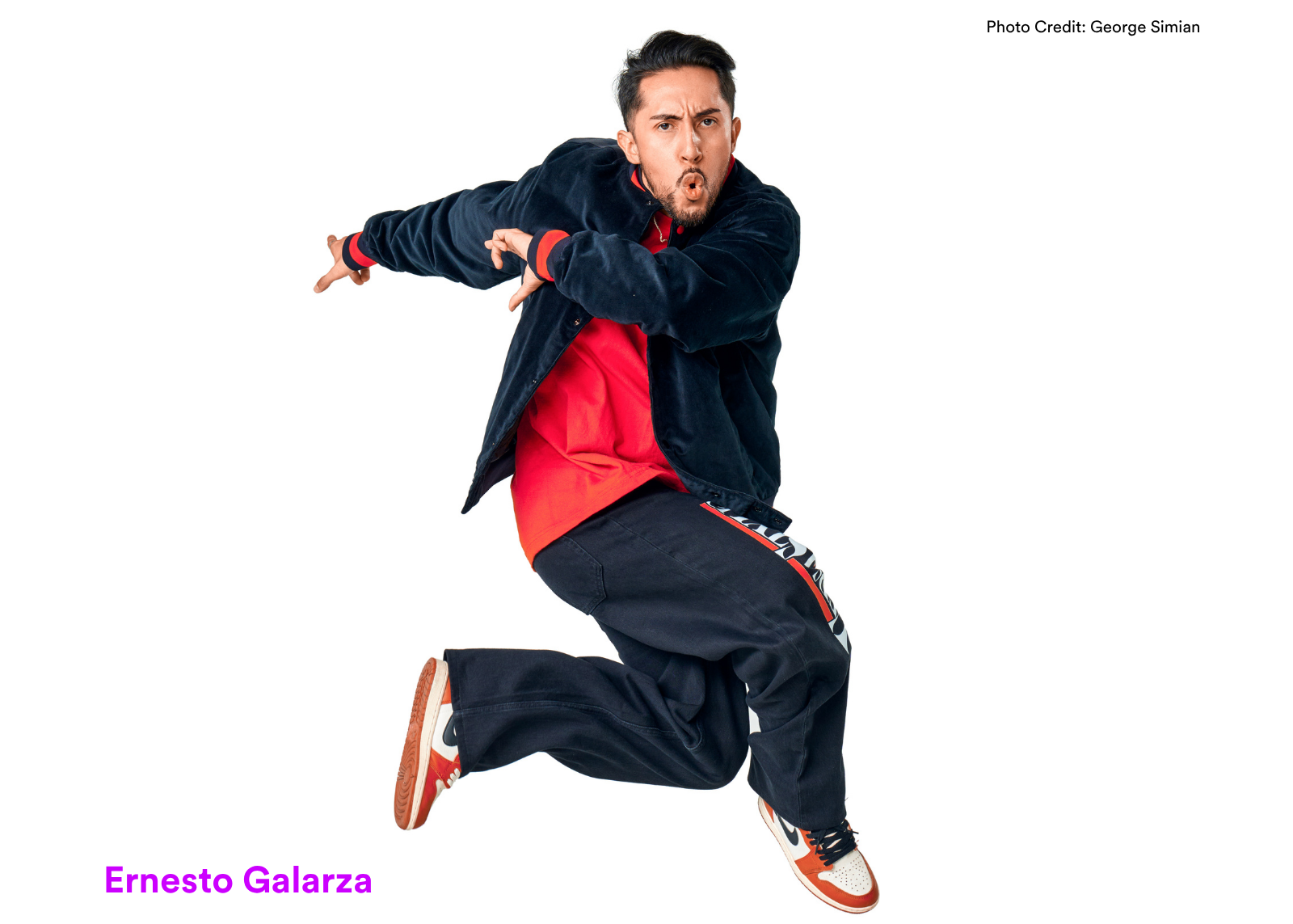 Dancer Ernesto Galarza leaps mid-air with arms crossed in a sharp pose, mouth open, wearing a navy jacket and red hoodie. Photo: George Simian.