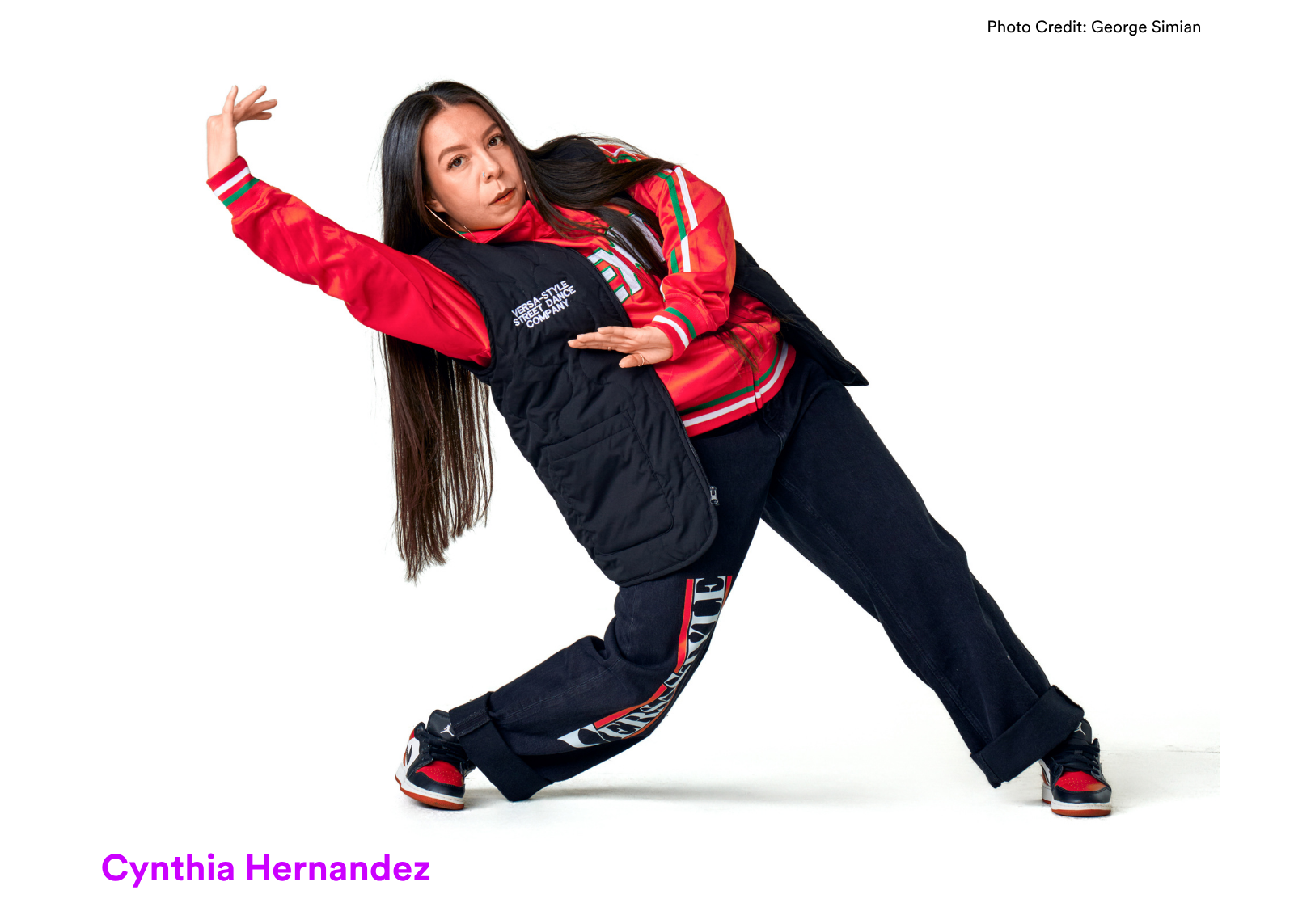 Dancer Cynthia Hernandez holds a deep side lunge with one arm extended, wearing a red satin bomber jacket and black vest. Photo: George Simian.