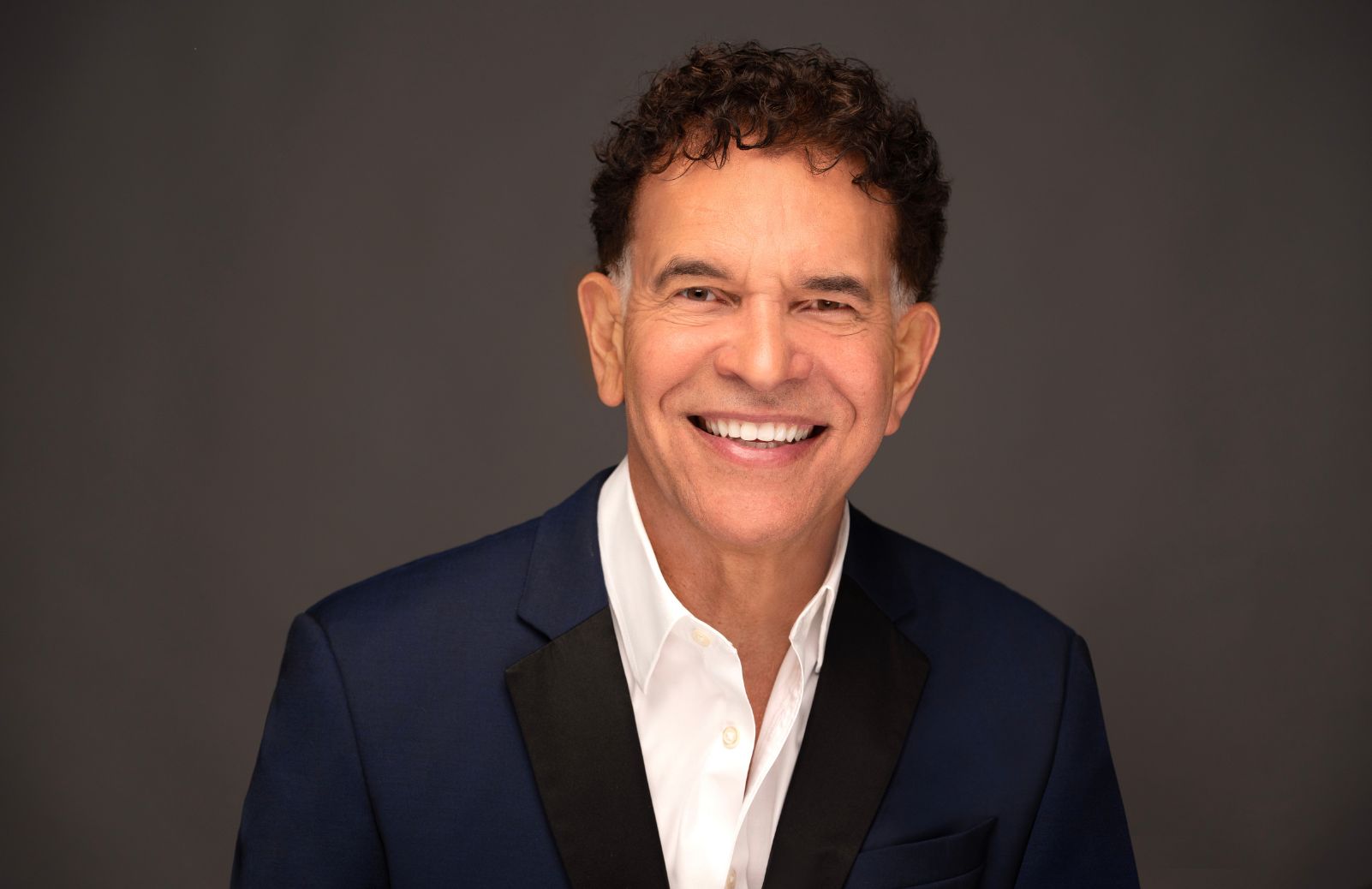 A smiling man in a navy blazer against a gray background.