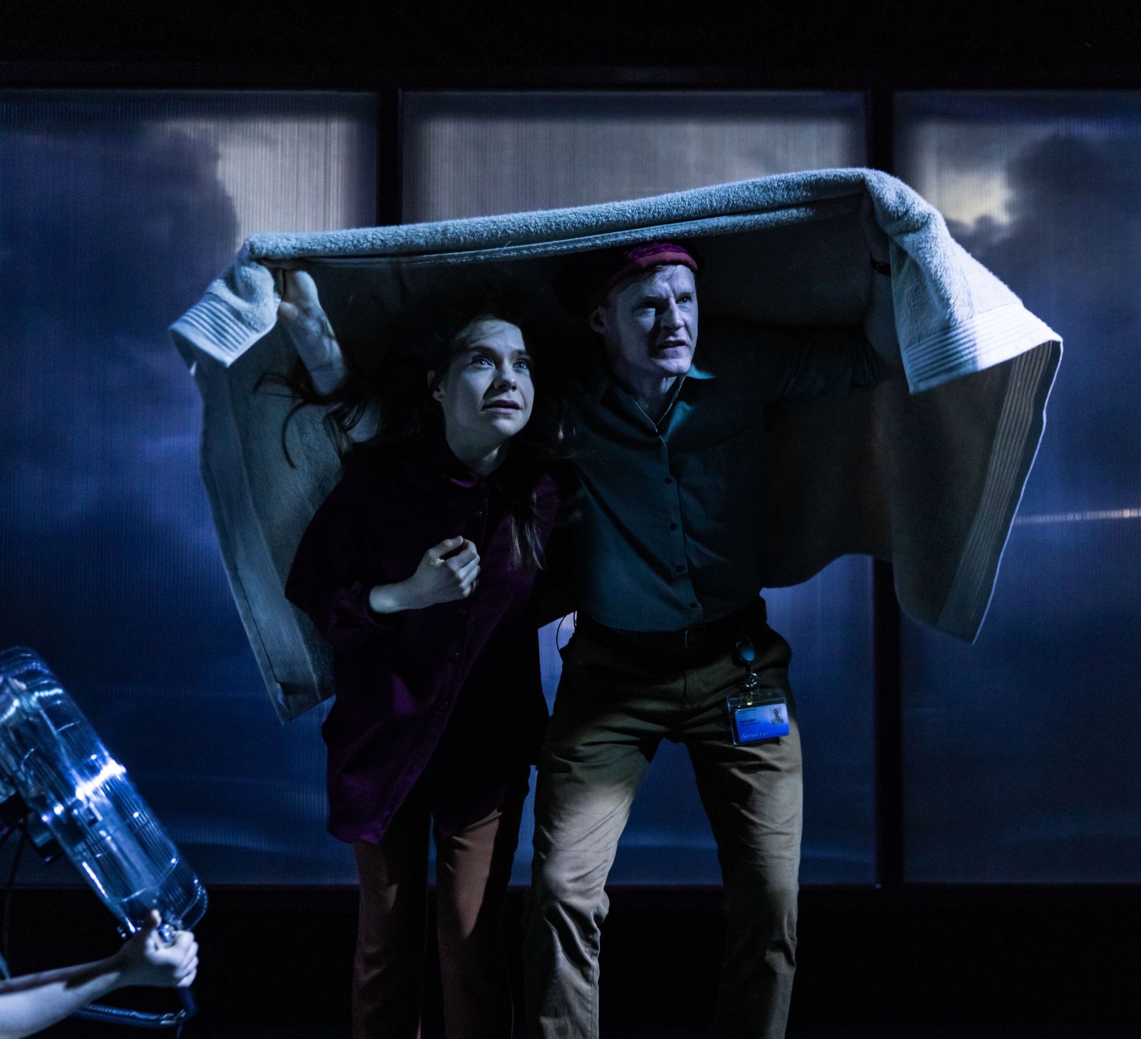 Two performers huddle under a blanket on a stormy, dramatically lit stage.