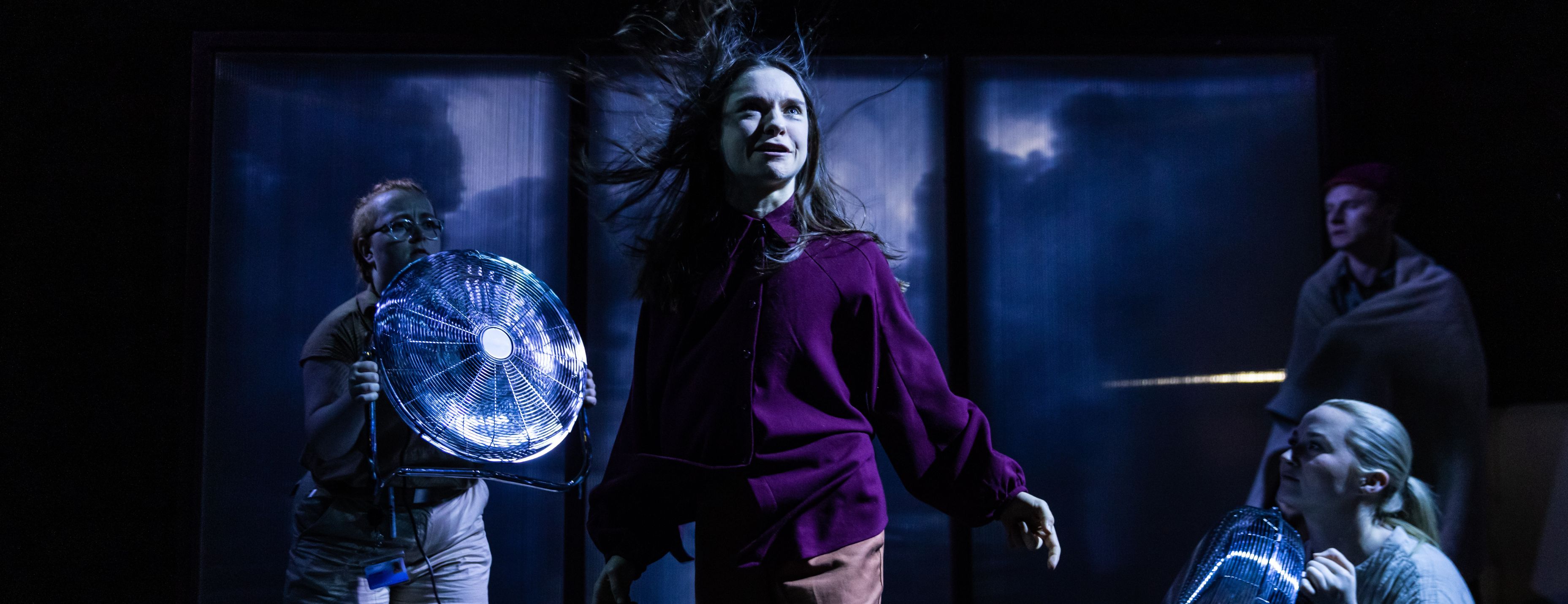 A performer's hair whips in the wind on a dark stage as cast members hold electric fans around her.