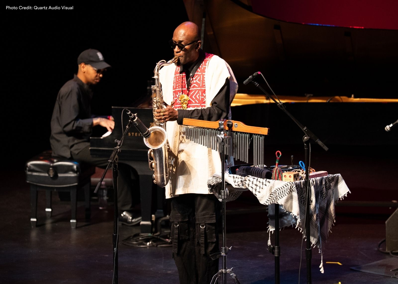 A saxophonist wearing sunglasses and a white embroidered tunic performs on stage next to a table of percussion instruments, with a pianist in the background.