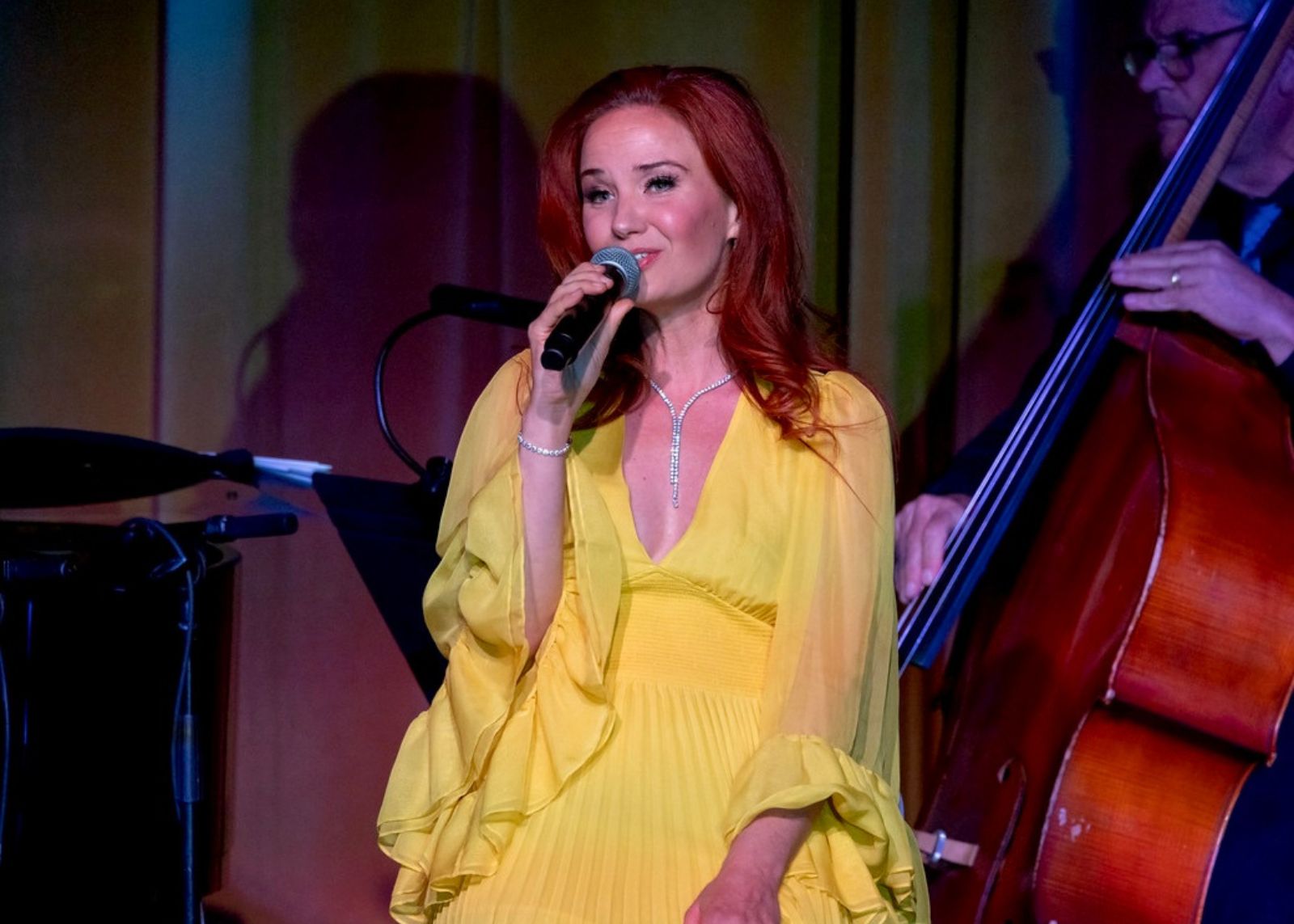 Singer in a flowing yellow gown performs with a handheld microphone, accompanied by a double bassist in a warm-lit venue.