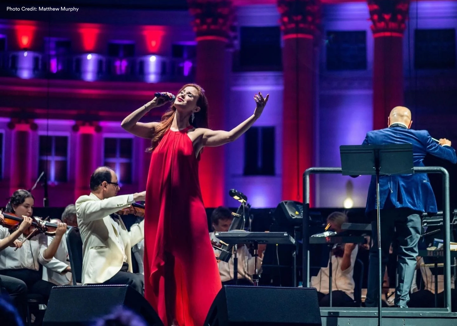 Singer in a flowing red gown performs with microphone in hand, arm extended, backed by a full orchestra under red and purple stage lighting.