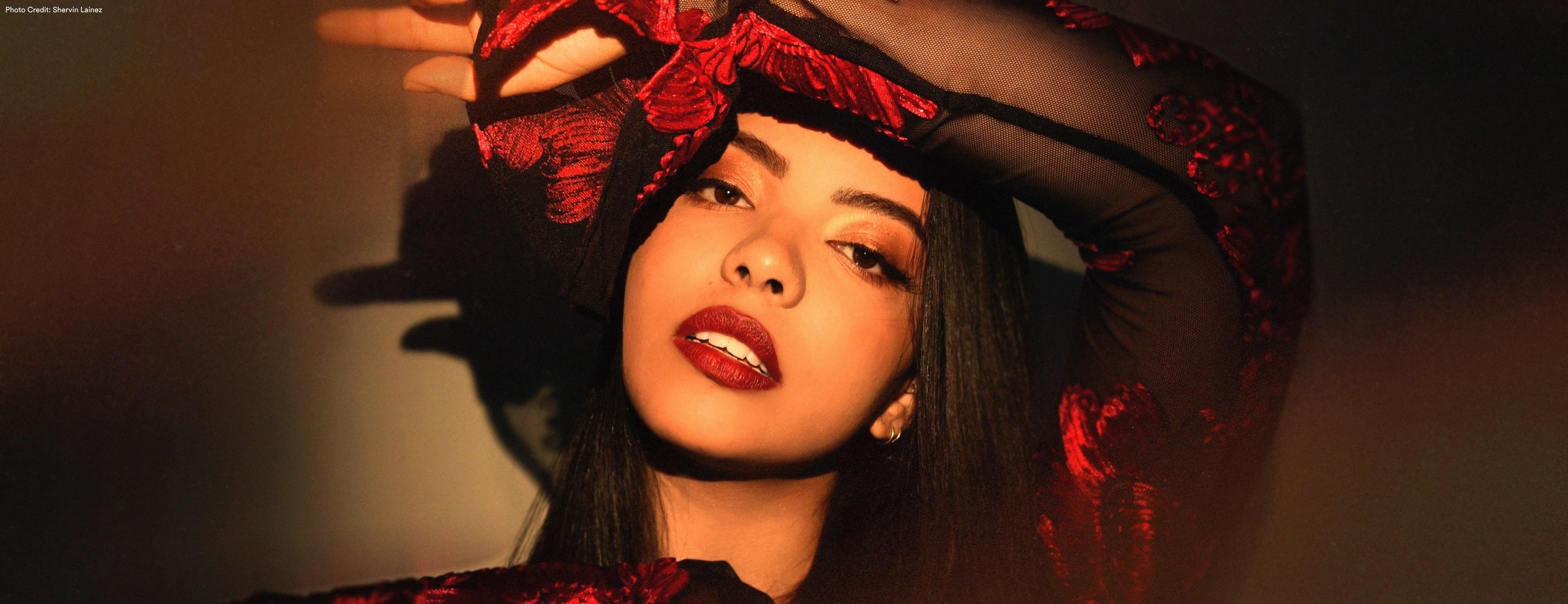 A woman gazes directly at the camera with one arm raised overhead, wearing a red lace top and matching headpiece in dramatic, moody lighting. Photo: Sharon Lavea.