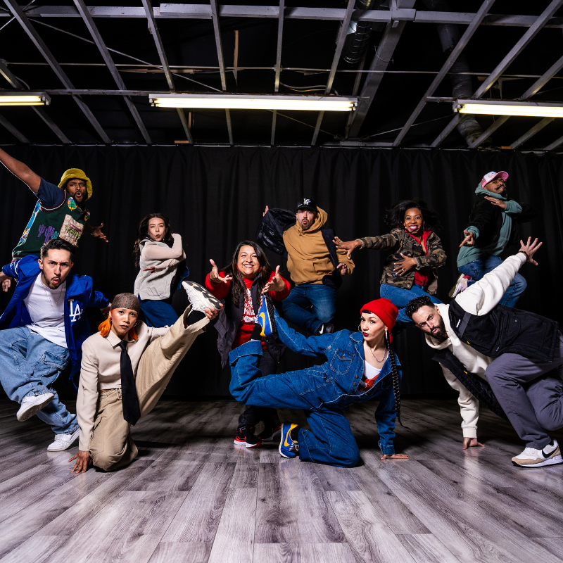 The full Versa-Style Dance Company ensemble strikes dynamic poses in a studio, some jumping and others low to the ground, lit dramatically against a black curtain backdrop. Photo: Timothy Salaz.