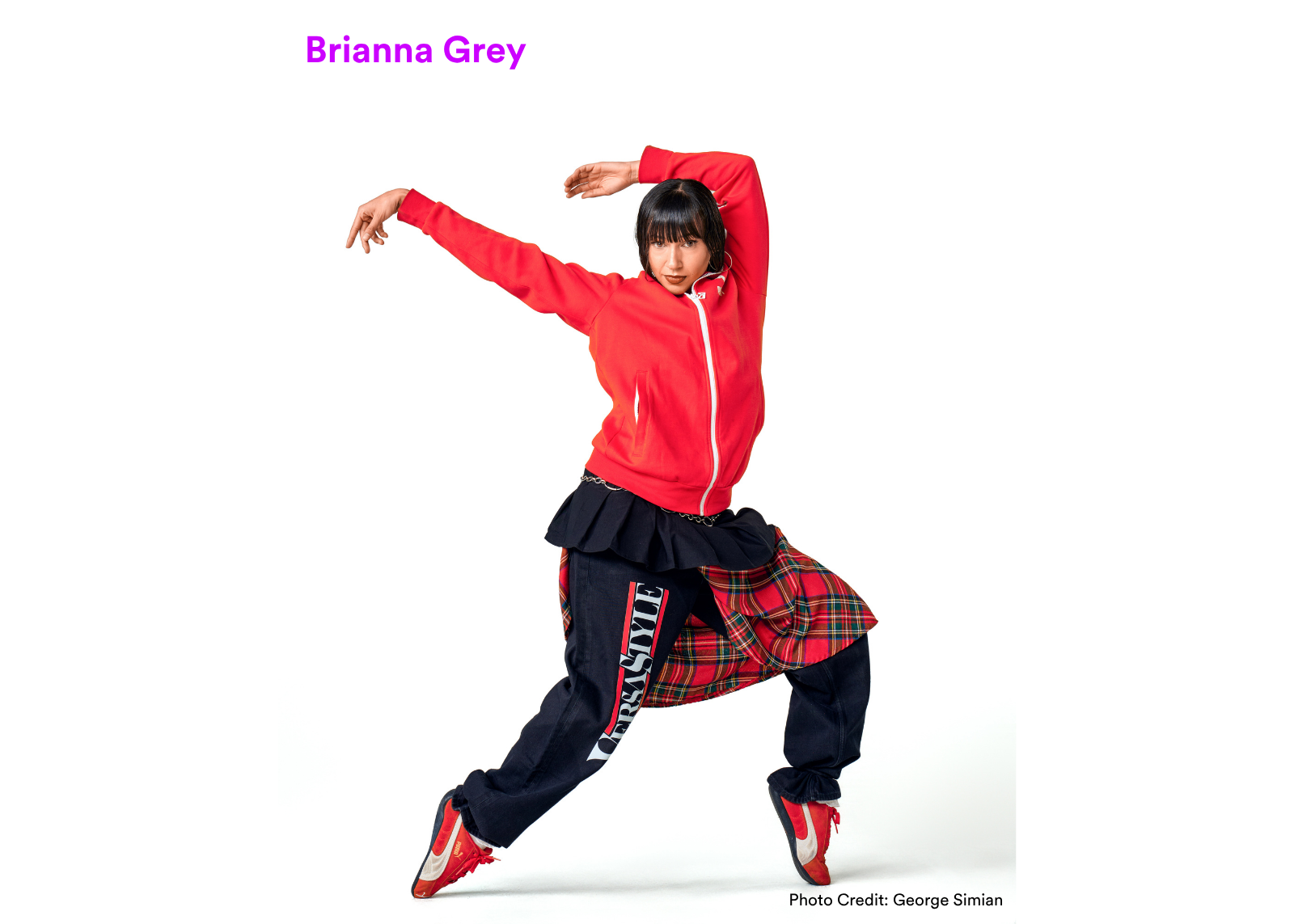 Dancer Brianna Grey holds a wide low stance with one arm arched overhead, wearing a red zip-up hoodie, black skirt, and plaid wrap. Photo: George Simian.
