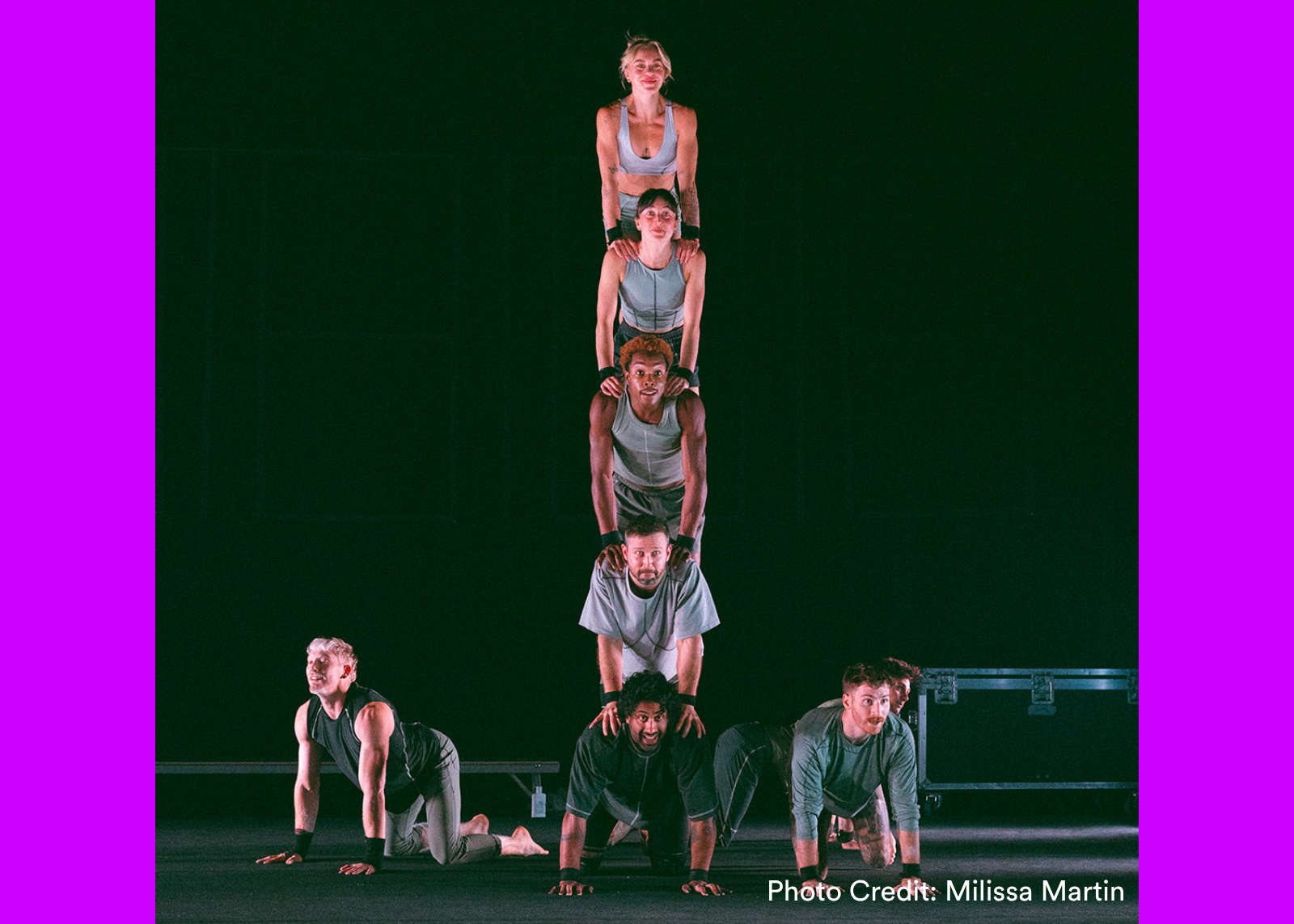 Six acrobats form a four-person-high human tower on a black stage, with three others positioned low on either side. (Photo Credit: Milissa Martin)