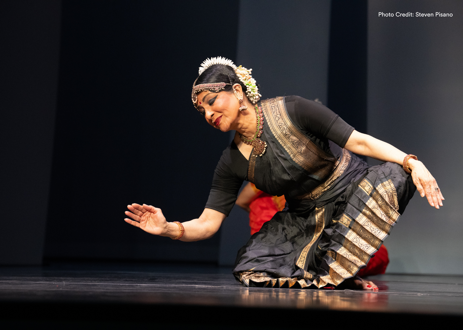 A dancer in a black and gold sari crouches low to the stage floor with arms extended. Photo: Steven Pisano.