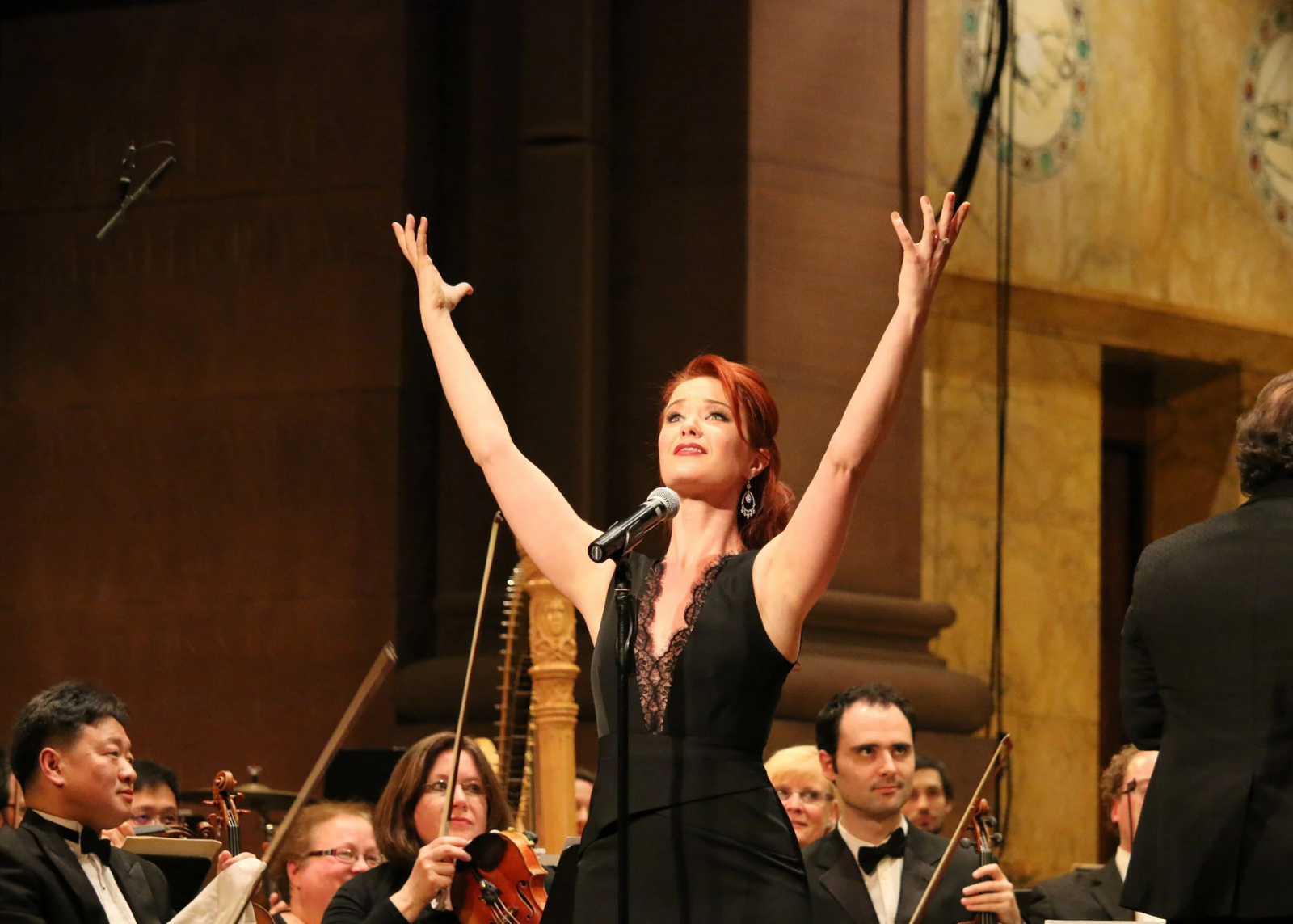 Singer with arms raised overhead performs before a symphony orchestra in a formal concert hall.