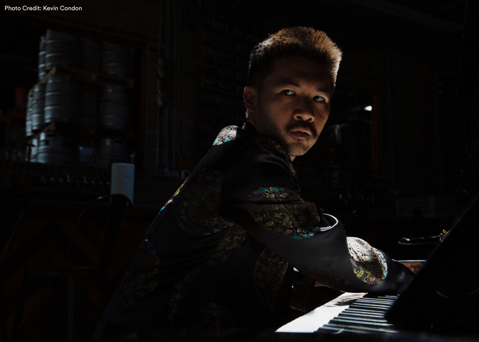 A pianist in a colorful patterned jacket looks back over his shoulder at the camera in dramatic low lighting. Photo: Kevin Condon.