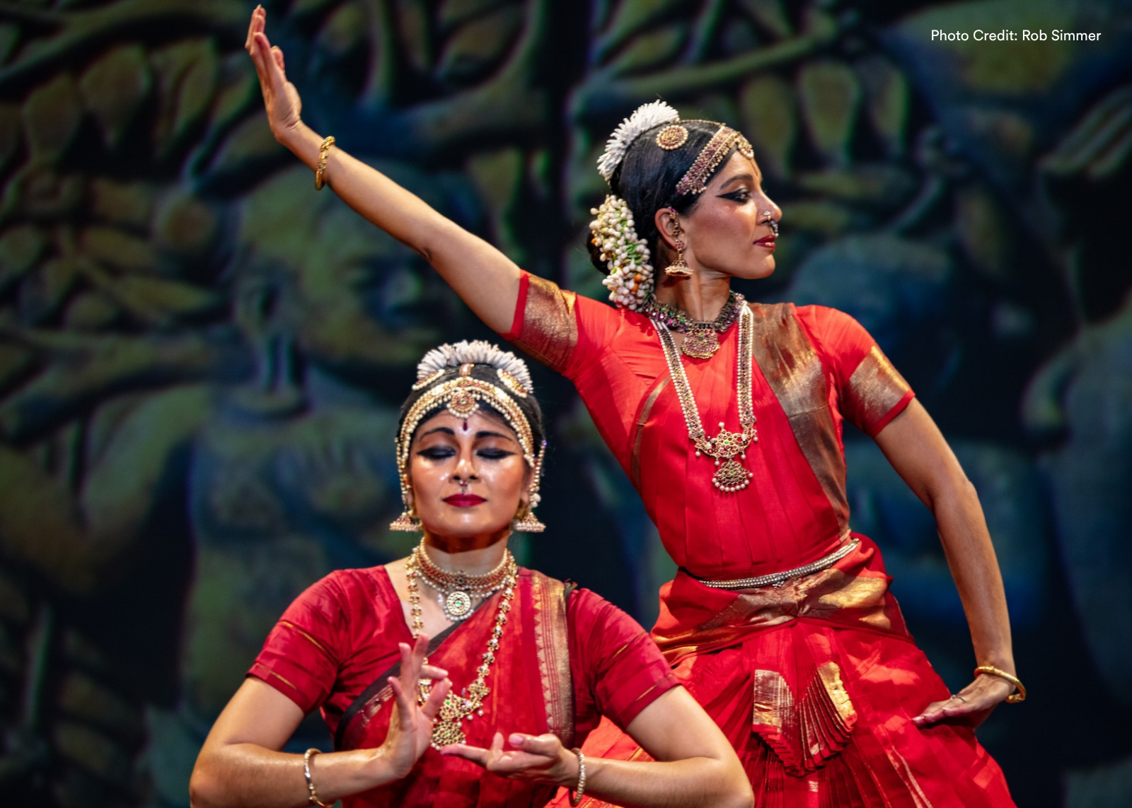 Two classical Indian dancers in red silk saris and traditional jewelry perform against a painted forest backdrop. Photo: Rob Simmer.