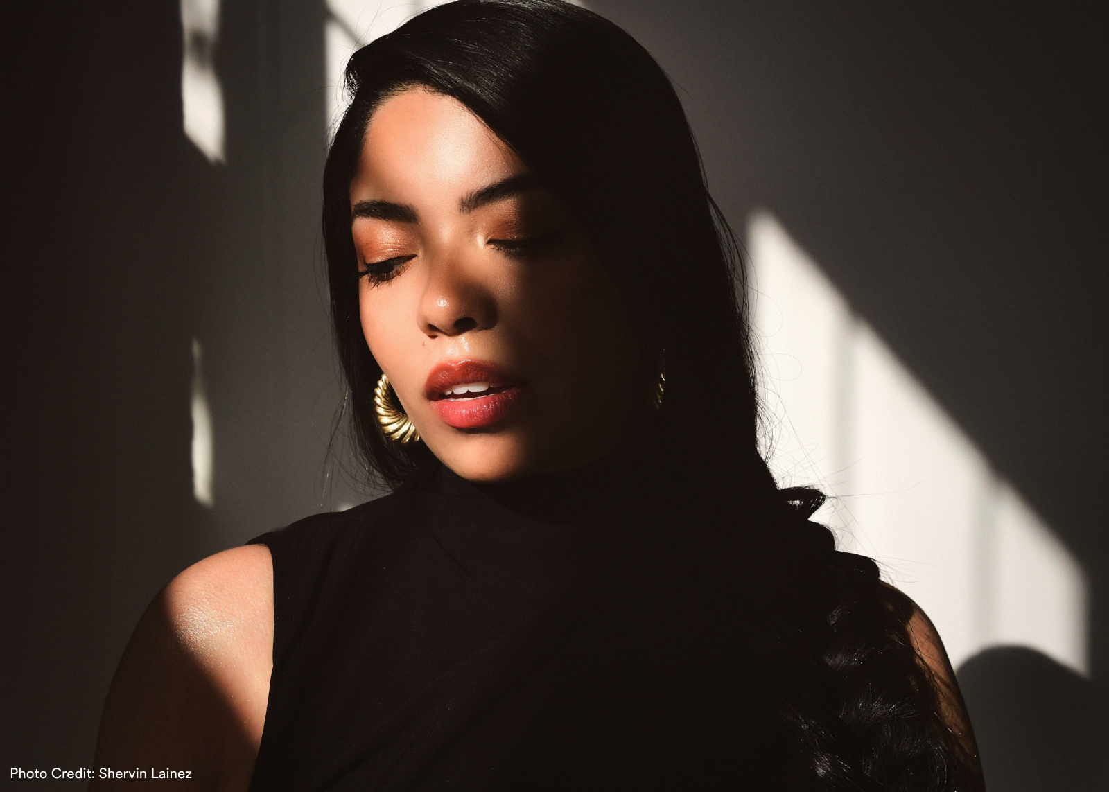 A woman with eyes closed tilts her head downward in dramatic shadow-play lighting, wearing a black top and gold hoop earrings. Photo: Shervin Lainez.